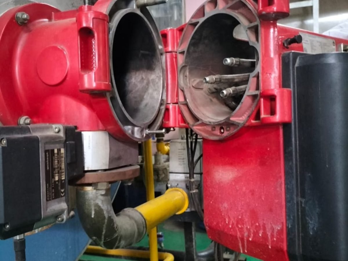 Rumah burner diayun terbuka — kepala pembakar, nosel, dan elektroda terlihat, beserta jelaga yang menempel di dalam blast tube